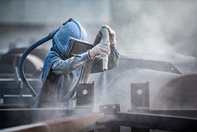 A worker in protective gear uses a hose to sandblast metal structures, creating a cloud of dust in an industrial setting.