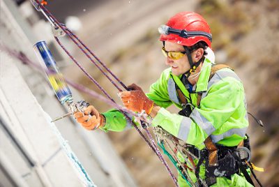 A worker in safety gear and harness paints a building exterior while suspended by ropes.