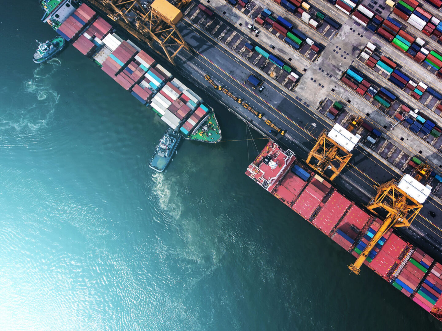 Aerial view of cargo ships docked at a port, with shipping containers stacked on the dock and cranes for loading and unloading.