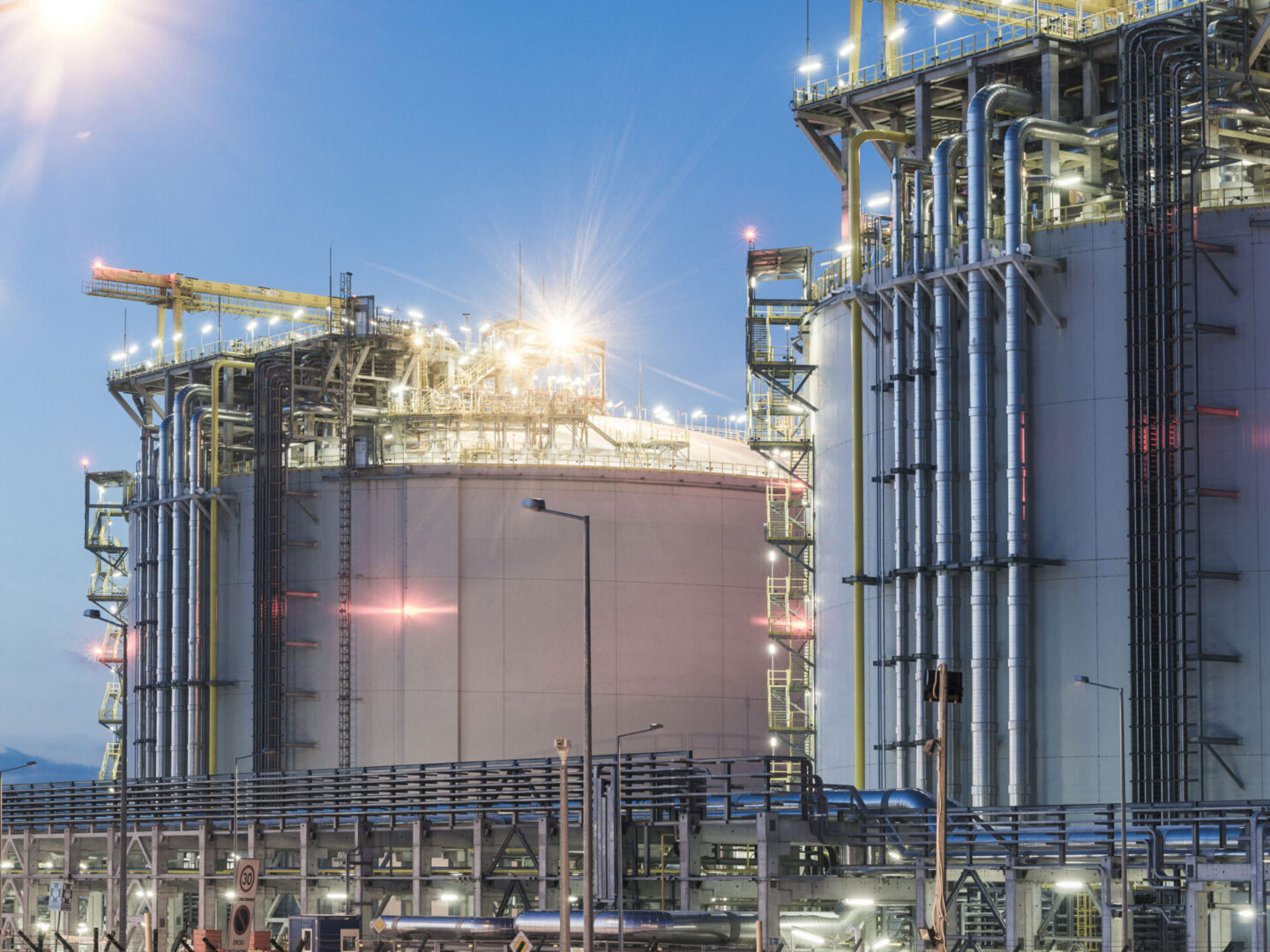 Large industrial storage tanks and metal piping at a refinery or chemical plant, illuminated by bright lights during twilight.